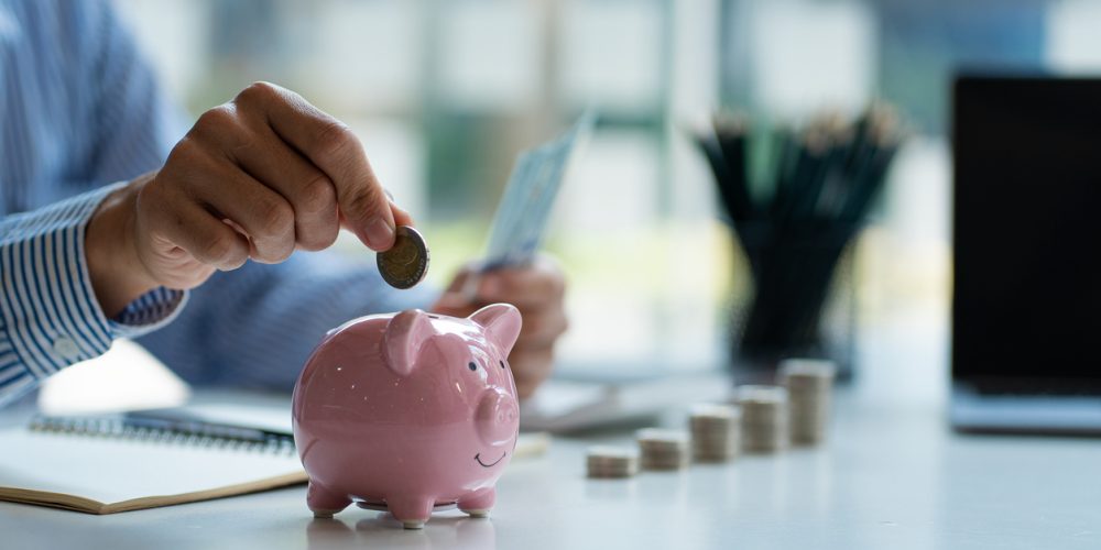 Close up image of a man's hand in an officer placing a quarter into a small pink piggy bank. In the background, the man's left hand is holding money.