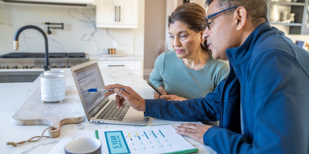 Older Couple sitting in kitchen in front of a computer and financial paperwork. The man is pointing at the computer screen with a pen while the wife looks at the computer screen