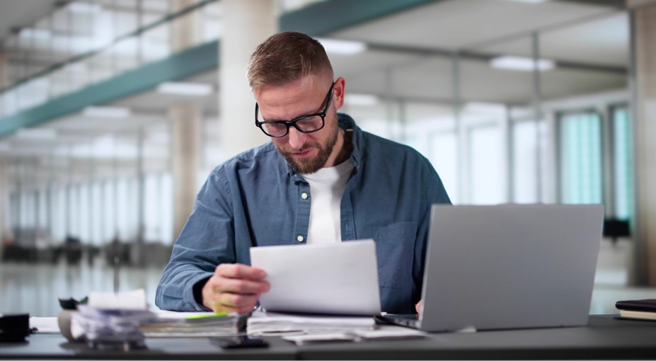 Business owner reviewing financial documents at his desk with a laptop, comparing paperwork for his small business