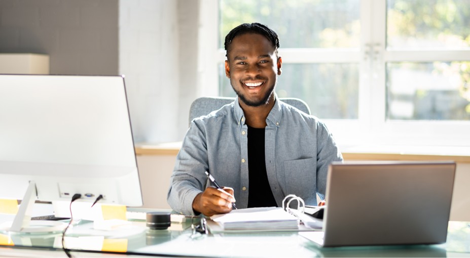 Smiling small business owner writing notes at his desk with a desktop computer and laptop, managing his business finances
