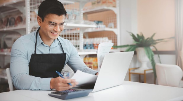 Smiling man in an apron using a laptop and calculator in a shop