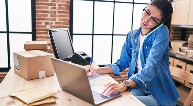 Young woman business owner talking on the phone while working on a laptop and taking notes at a desk with shipping boxes in a warehouse workspace